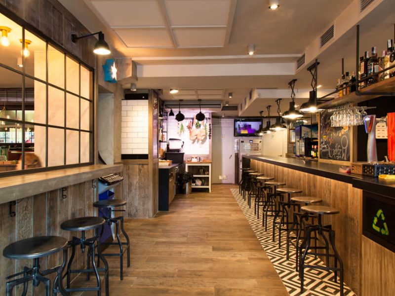 Interior view of a commercial establishment with durable laminate flooring in an oak finish, arranged in a straight plank pattern. The setting includes a bar area with stools, industrial-style lighting, and a mix of rustic and contemporary decor elements.