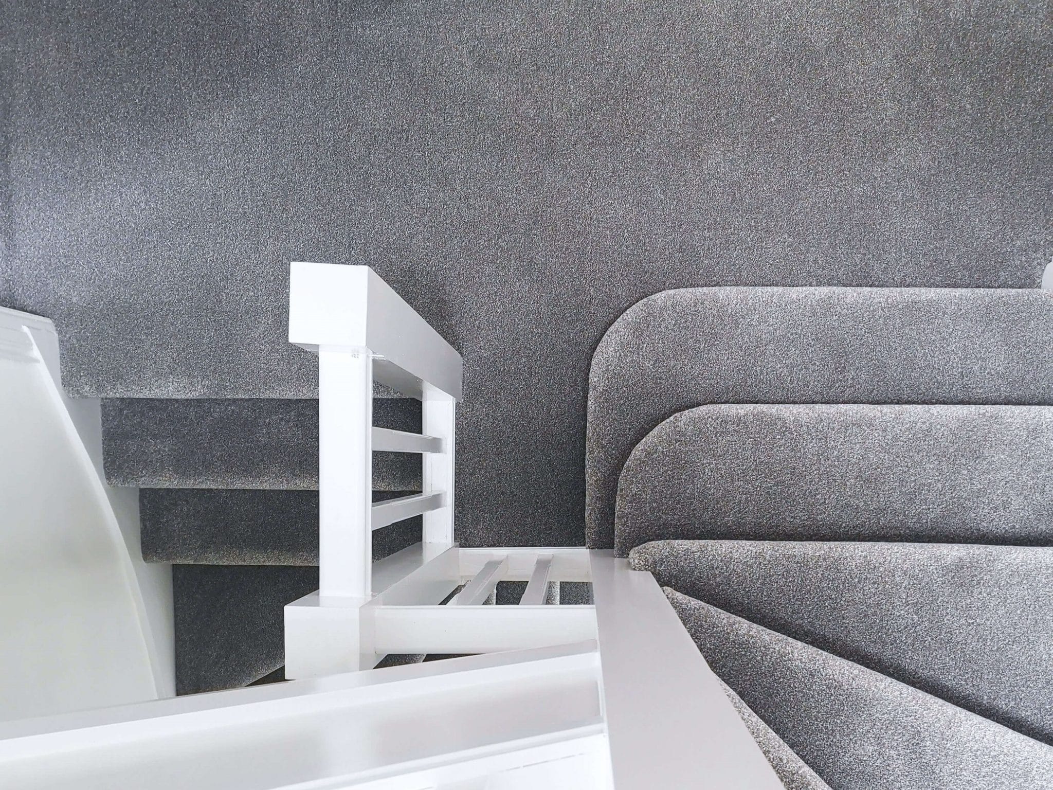 Image of a modern interior stairway with white balusters and handrail alongside a curved grey carpeted staircase. The walls are textured with a speckled grey finish, complementing the carpet's colour and texture
