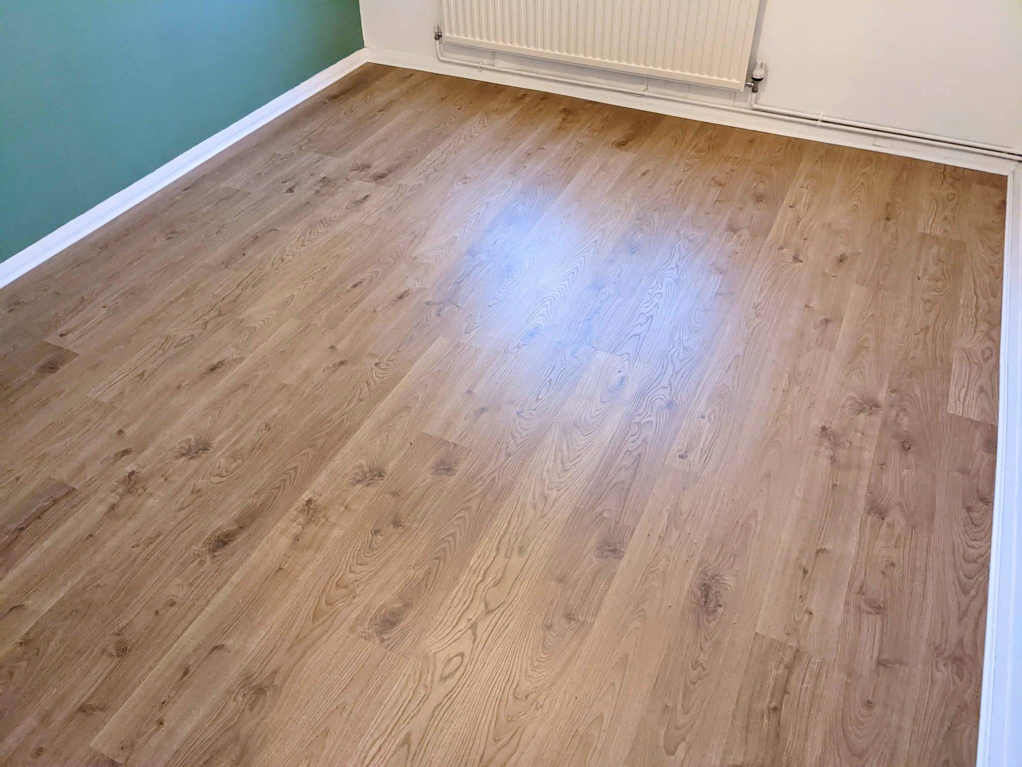 Image of a room with QuickStep laminate flooring in a light oak finish, showcasing the planks' detailed wood grain pattern. The room features green walls, white skirting, and a white radiator, highlighting the warm tones of the laminate.